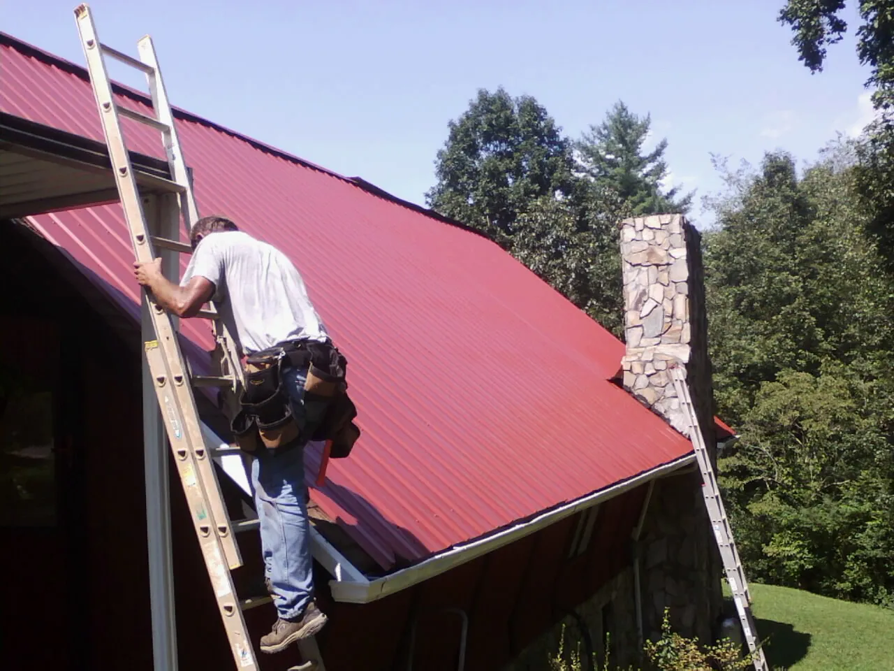 Roofer on a ladder beside a red metal roof and stone chimney — Soffit & Fascia Repair by Roofing Brothers in Mount Savage